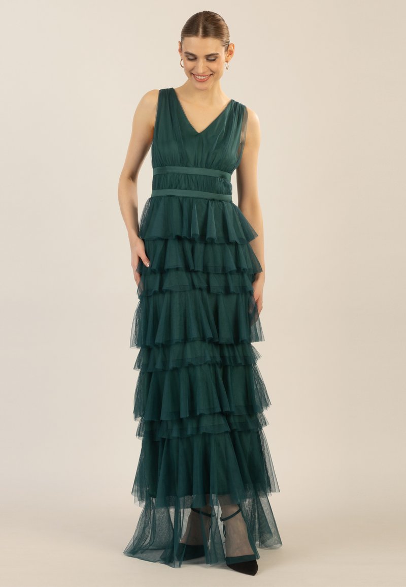 Langes, gestuftes Abendkleid aus tiefgrüner Tüll, mit V-Ausschnitt, Taillengürtel und geschichteten Rüschen, kombiniert mit schwarzen High Heels.