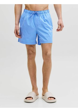 Bărbat purtând pantaloni scurți de baie albastru deschis și sandale bej, stând în picioare pe un fundal simplu și deschis.