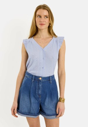 Mujer con cabello rubio largo que lleva una blusa azul claro sin mangas con ojales y shorts vaqueros de cintura alta con los bajos remangados, pulsera de oro en la muñeca.