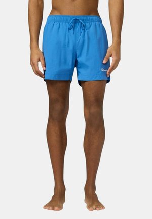 Uomo che indossa pantaloncini da bagno azzurri vivaci con coulisse, in piedi a piedi nudi su uno sfondo chiaro e uniforme.