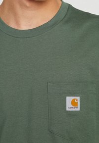Man wearing olive green Carhartt T-shirt with a chest pocket featuring the Carhartt logo patch.