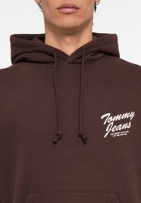 Bruine hoodie van zacht materiaal, met een voorzak en trekkoorden. Opvallende witte bedrukte tekst op de linkerborst.