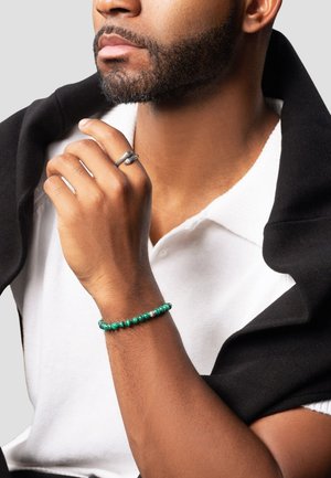 Grøn perlesmykke med sølvdetaljer på venstre håndled; modellen bærer en hvid skjorte med krave og en sort jakke over skulderen.