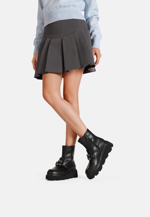 Stivali neri in pelle fino alla caviglia con suola spessa, dettaglio con fibbia e texture liscia, abbinati a una gonna grigia plissettata e un maglione azzurro chiaro.