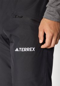 Svarta utomhusbyxor med en elegant design, som har en vit "TERREX"-logotyp på låret och en fickdetalj med dragkedja på sidan.