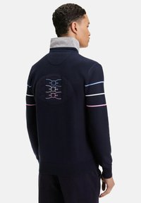 Pull marin avec un col montant et des poignets côtelés. Présente un logo texturé au dos et des rayures colorées sur les manches en bleu, blanc et rose.