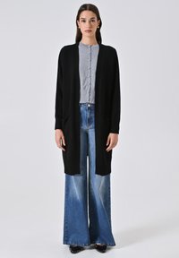 Frau steht in einem langen schwarzen Cardigan, grauem Knopf-Oberteil, weit geschnittenen blauen Jeans, schwarzen Schuhen und goldenen Ohrringen vor weißem Hintergrund.