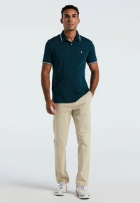 Teal poloshirt met een witte gestreepte kraag, gecombineerd met beige broek en witte sneakers. Het shirt heeft een subtiele logo op de borst.