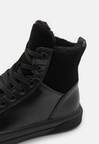 Adidași negri cu prindere înaltă, având un exterior din piele lacuită și suede, șireturi rotunde și o căptușeală moale neagră pentru un plus de confort.