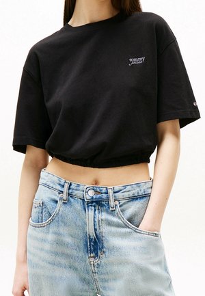 Kvinde iført en sort cropped Tommy Jeans T-shirt og lyseblå højtaljede denimjeans med den ene hånd i lommen.