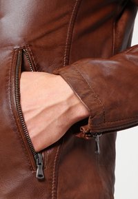 Brun läderjacka med en närbild av en hand i en dragkedjeficka, med en mjuk yta och kontrasterande metalldetaljer på dragkedjan.