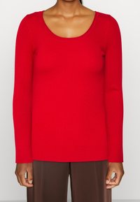 Rotes, geripptes Langarmoberteil mit U-Boot-Ausschnitt; figurbetontes Design und glatte Textur; kombiniert mit braunen Hose mit weitem Bein.