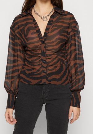 Hun har en gennemsigtig, langærmet, knapbluse med brune og sorte tigerstriber og sorte bukser på, accessoriseret med en sølvkæde halskæde.