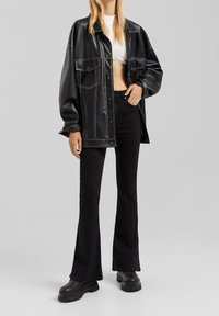 Musta tekonahkakangastakki, jossa on valkoinen ommel, kahdella etutaskulla ja rento istuvuus, yhdistettynä liehuviin mustiin housuihin ja paksupohjaisiin kenkiin.