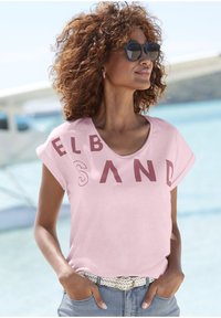Roze T-shirt met korte mouwen en ronde hals, met gedurfde kastanjebruine letters die "ELB SAND" spellen. Dit is gecombineerd met lichte spijkerbroekshorts en een gevlochten riem.