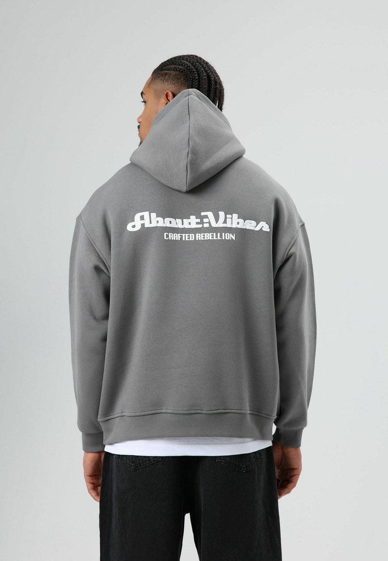 Grijze oversized hoodie met een grote witte grafische opdruk op de achterkant met de tekst "About: Vibes" en "CRAFTED REBELLION." Zacht materiaal, hooded ontwerp.