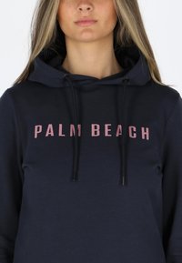 Felpa a cappuccio blu navy realizzata in tessuto morbido con la scritta rosa "PALM BEACH". Include i lacci e ha un design dalla vestibilità rilassata.