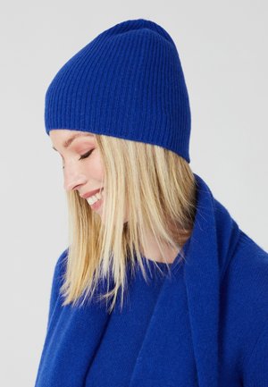 Blauwe gebreide beanie met ribbelstructuur, ronde kroon en zachte stof. Gecombineerd met een blauwe trui en sjaal, wat het monochrome ontwerp benadrukt.