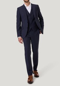 Granatowy garnitur szyty na miarę z klapami typu notch, kamizelka zapinana na trzy guziki oraz dopasowane spodnie. Brązowe skórzane buty typu double monk strap.