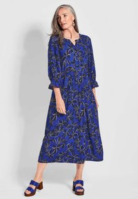 Blaues Blumenkleid mit langen Ärmeln, Knopfleiste, lockerer Passform und Rüschensäumen; kombiniert mit blauen Blockabsatzsandalen.