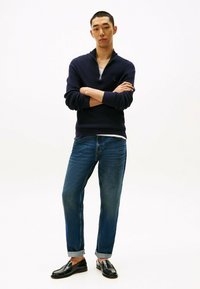 Pull zippé bleu marine, jeans en denim et mocassins noirs. Le pull présente un tricot texturé ; les jeans sont décolorés et retroussés aux chevilles.