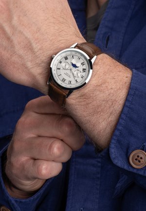 Montre en argent avec un bracelet en cuir marron, boîtier circulaire, marqueurs en chiffres romains, trois sous-cadrans et indicateur de phase de lune, portée au poignet.