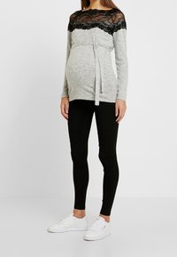 Haut de maternité gris avec encolure en dentelle noire, manches longues et détail à nouer sur le côté, associé à des leggings noirs et des baskets blanches.