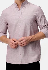 Chemise boutonnée de couleur violet clair avec une poche poitrine, manches longues, texture subtile et boutons roses, portée par une personne qui l'ajuste.
