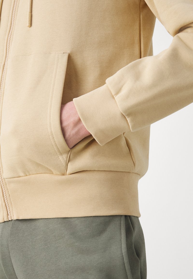Beige zip-up hoodie met een kangoeroezak, geribbelde manchetten en een zachte textuur, gecombineerd met donkergroene trainingsbroek.