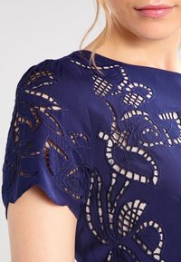 Navy blue dress with intricate floral embroidery and sheer cutouts on sleeves, made of smooth satin fabric. Fitted design with scalloped edges.