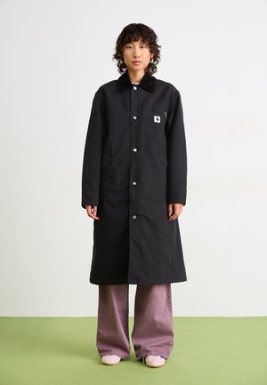 Personne avec des cheveux courts et bouclés portant un long manteau noir sur un pantalon large violet et des chaussures rose clair, se tenant sur un sol vert.