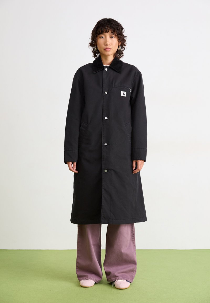 Personne avec des cheveux courts et bouclés portant un long manteau noir sur un pantalon large violet et des chaussures rose clair, se tenant sur un sol vert.