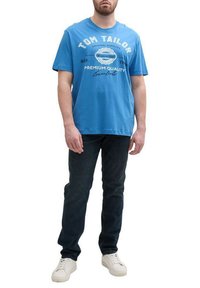 T-shirt en coton bleu avec un imprimé graphique portant le texte "TOM TAILOR". Associé à un jean en denim foncé et des baskets blanches.