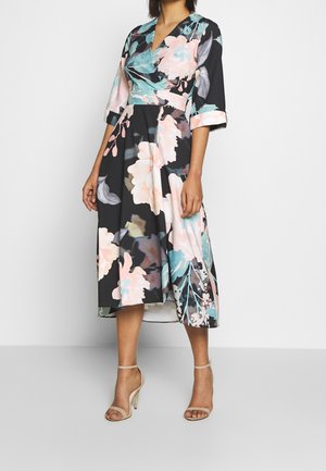 Femme portant une robe midi noire à imprimé floral rose et bleu, manches trois-quarts, et sandales à talons hauts beiges.