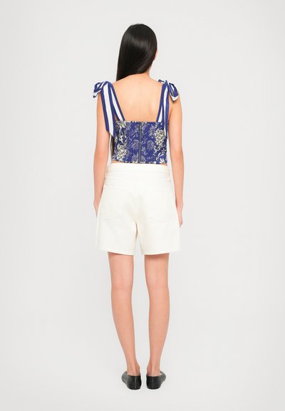 Woman with long dark hair wearing a blue floral crop top with striped tie straps and white high-waisted shorts, standing with her back to the camera.