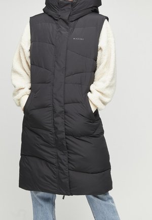 Long manteau sans manches matelassé noir avec poches sur le devant, porté par-dessus un sweat en polaire crème et un jean bleu clair.
