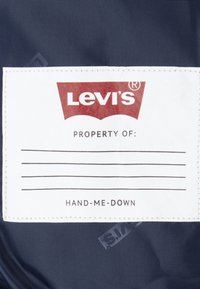 Étiquette Levi's sur tissu bleu marine ; présente une étiquette rectangulaire blanche avec "PROPERTY OF," des lignes vides, et "HAND-ME-DOWN" en texte noir gras.