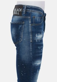 Blauwe spijkerbroek van denim met een verouderde look, distressed details en een zwart merkembleem op de tailleband. Voorzien van vijf zakken en een slim fit.