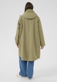 Personne portant un long manteau vert olive avec une capuche, un jean large bleu et des baskets blanches, vue de dos sur un fond uni.