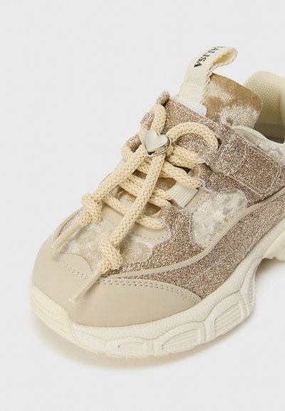 Beige Sneaker mit Glitzerdetails, dicke Sohle, cremefarbene Schnürsenkel, die mit einem silbernen herzförmigen Charm gebunden sind, auf weißem Hintergrund.