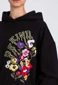Felpa con cappuccio nera con ricamo floreale colorato in tonalità di rosa, rosso, giallo e viola, accompagnata dal testo "BE KIND" in giallo.