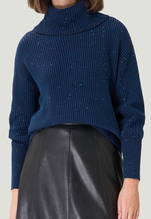 Femme portant un pull à col roulé côtelé bleu marine avec de petites paillettes et une jupe en cuir noir taille haute.