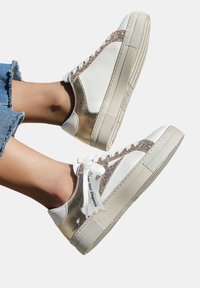 Baskets blanches avec des accents dorés brillants, semelle en caoutchouc texturée et surface lisse. Comprend un design à lacets et une étiquette imprimée.