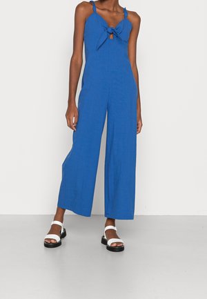 Vrouw draagt een mouwloze blauwe jumpsuit met een knoopdetail aan de voorkant en wijde pijpen, gecombineerd met witte plateauzandalen.