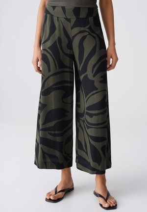 Pantalon à jambes larges avec imprimé floral abstrait en vert olive et noir, porté avec des tongs noires sur fond uni.