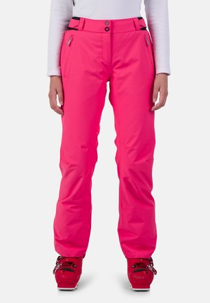 Pink ski pants with a straight cut, featuring a zippered pocket and adjustable waistband, paired with red ski boots.