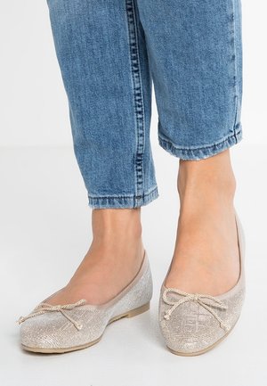Fødder iført glitrende beige ballerinasko med sløjfeaccenter kombineret med bukser i blå denim med opsmøg på en hvid gulv.