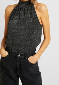 Haut noir à col licou avec une subtilité scintillante, présentant un design plissé. Associé à un jean denim foncé taille haute, les mains dans les poches.