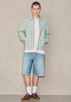 Young man standing wearing green plaid zip jacket, white T-shirt, light blue denim shorts, white socks, and brown boat shoes against beige wall.