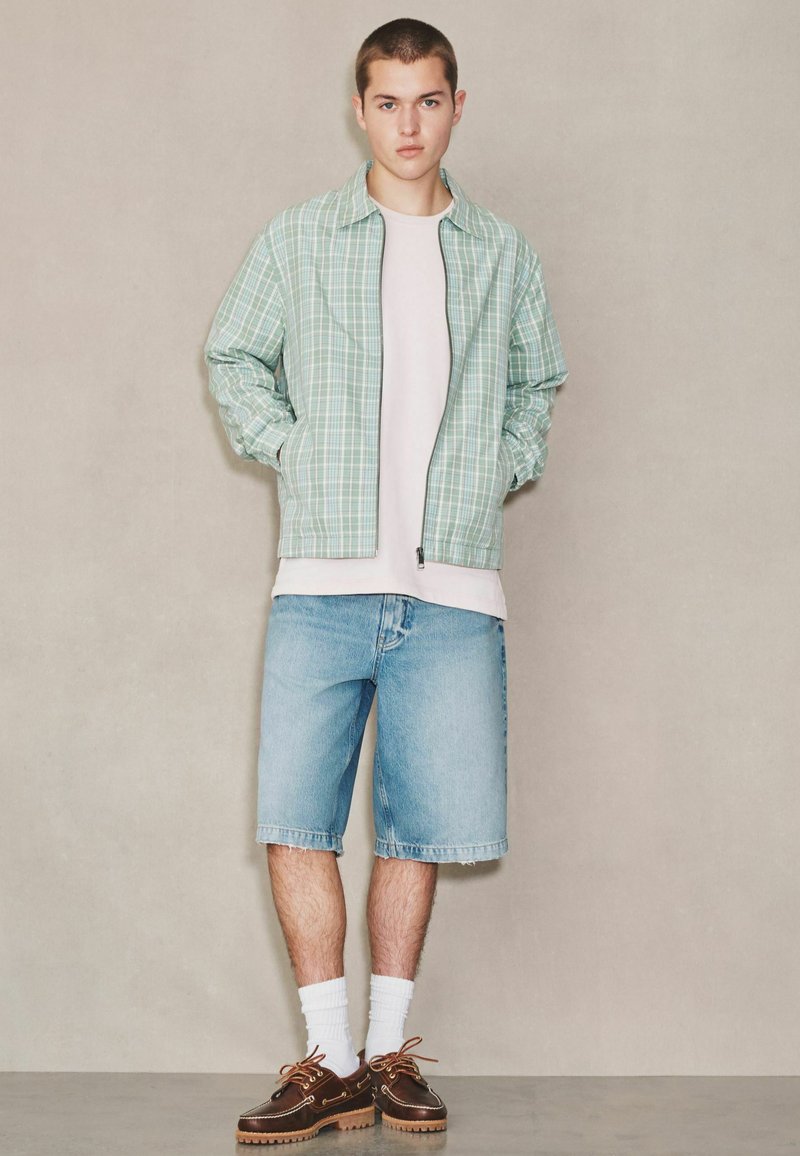 Jeune homme debout portant une veste zippée à carreaux verts, un t-shirt blanc, un short en jean bleu clair, des chaussettes blanches et des chaussures bateau marron, devant un mur beige.